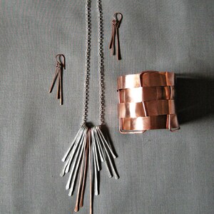 May include: A jewelry set featuring a silver chain necklace with a fan-shaped pendant, copper and silver earrings, and a copper cuff bracelet. The jewelry is displayed on a gray background.