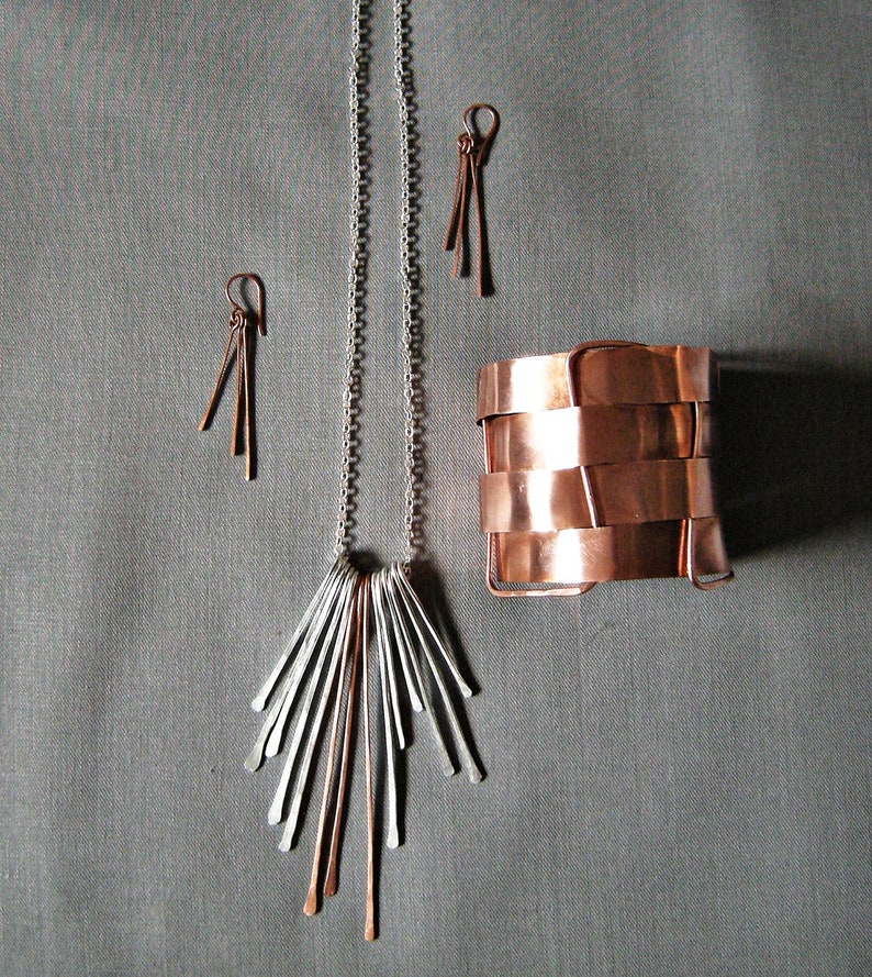 May include: A jewelry set featuring a silver chain necklace with a fan-shaped pendant, copper and silver earrings, and a copper cuff bracelet. The jewelry is displayed on a gray background.