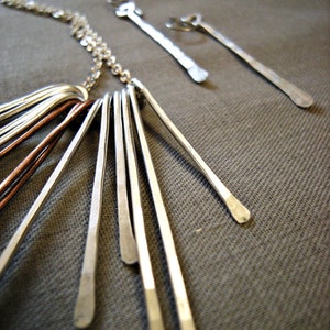 May include: A silver necklace with multiple dangling bars, some copper-colored, and matching earrings. The jewelry is displayed on a gray textured surface. The necklace has a delicate chain and the bars have a hammered texture.