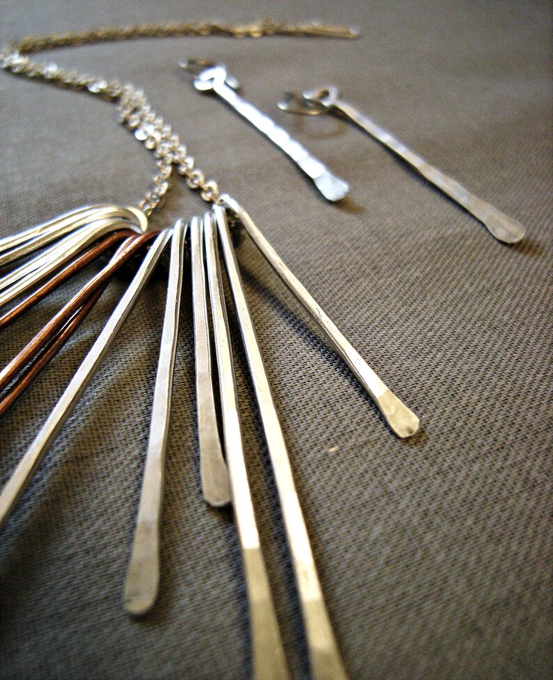 May include: A silver necklace with multiple dangling bars, some copper-colored, and matching earrings. The jewelry is displayed on a gray textured surface. The necklace has a delicate chain and the bars have a hammered texture.