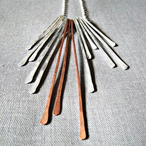 May include: A silver and copper-toned pendant necklace. The necklace features a series of silver and copper-colored bars radiating from a central point. The bars are arranged in a fan-like pattern, creating a modern and geometric design. The necklace is displayed on a gray fabric surface.