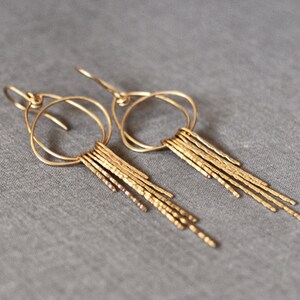 May include: Gold dangle earrings with a circle design and a fringe of textured metal strands.