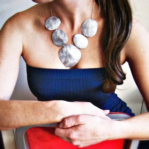 May include: A silver necklace with a layered design of round, textured pendants. The necklace is worn by a person with long brown hair and a blue strapless top.