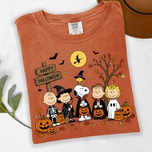 May include: A rust-colored t-shirt featuring a graphic of Peanuts characters in Halloween costumes. The design includes Snoopy, Charlie Brown, and friends with pumpkins, a wooden sign reading Happy Halloween, a witch on a moon, and bats.