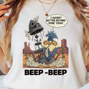 May include: Cream-colored t-shirt featuring a cartoon roadrunner character with a desert landscape backdrop. The design includes the text "I HAVEN'T GOTTEN NOTHING DONE TODAY" in a speech bubble and "BEEP - BEEP" at the bottom.
