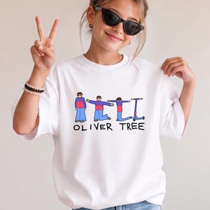 May include: A white short-sleeved t-shirt featuring a graphic print of a cartoon figure in a red and blue jacket with blue pants, shown in various poses alongside a scooter, with the text OLIVER TREE printed in black capital letters below.