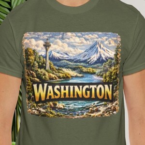 May include: Olive green t-shirt with a landscape design and the word "WASHINGTON" in gold. The design features a river, mountains, and a tower, reminiscent of a vintage travel poster.