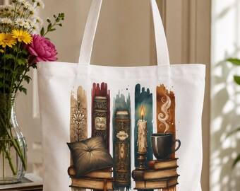 May include: A white canvas tote bag featuring a colorful graphic print of stacked books, a lit candle, a steaming mug, and a small pillow. The text "Just one more chapter" is printed in black at the bottom center of the bag, which has two white handles.