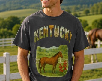 May include: A charcoal gray short-sleeve t-shirt featuring the word KENTUCKY in large block letters filled with a landscape scene. Below the text is a rectangular graphic of a brown horse standing in a green pasture with red flowers.