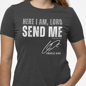 以下が含まれることがあります： 「HERE I AM, LORD SEND ME」の白文字がプリントされた濃いグレーのTシャツ。テキストの下には「Isaiah 6:8」と署名があります。Tシャツはクルーネックで半袖です。