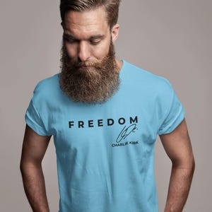 以下が含まれることがあります： 黒字で「FREEDOM」の文字がプリントされた水色のTシャツ。サインと「CHARLIE KIRK」の名前も含まれています。Tシャツは半袖です。