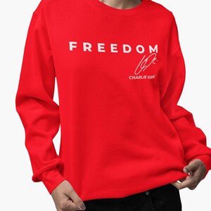 以下が含まれることがあります： 胸に白い文字で「FREEDOM」とプリントされた鮮やかな赤いスウェットシャツ。その下に白いサインと「Charlie Kirk」の名前があります。スウェットシャツは無地で、長袖です。