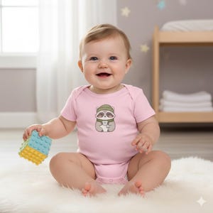 Op de afbeelding: Een lachende baby draagt een lichtroze romper met een wasbeer-afbeelding. De wasbeer draagt een groene muts en sjaal. De baby houdt een blauw en geel speelgoedblok vast. De romper is een babykledingstuk.