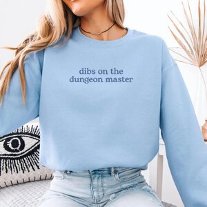 May include: A light blue sweatshirt with the text "dibs on the dungeon master" in a dark blue font. The long-sleeved crew neck sweatshirt is made of a soft material.