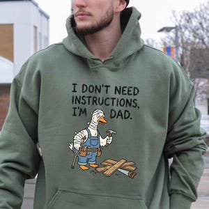 May include: Olive green hoodie with a front pocket and drawstring hood. The front features the text "I DON'T NEED INSTRUCTIONS, I'M DAD." below a cartoon duck wearing a bandage, overalls, and holding tools. The duck is standing on wooden planks.