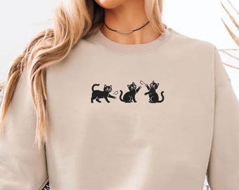 May include: Beige sweatshirt featuring a black graphic of three cats. The cats are in different poses, with one walking, one sitting, and one holding a small object. The sweatshirt has a crew neck and long sleeves.
