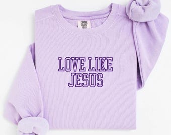 May include: A light purple sweatshirt with the text "LOVE LIKE JESUS" in a darker purple font. The sweatshirt has long sleeves with cuffed ends and a crew neck. The Comfort Colors brand tag is visible.