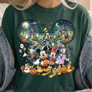 K&ouml;nnte beinhalten: Ein dunkelgr&uuml;nes T-Shirt mit einer gro&szlig;en Grafik von Zeichentrickfiguren in Halloween-Kost&uuml;men. Das Design ist in Form einer Maus-Kopf-Silhouette gehalten und zeigt ein Spukschloss, K&uuml;rbisse, Geister, Flederm&auml;use und Sterne in einer Nachtszene.
