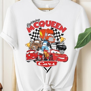 May include: White t-shirt featuring a colorful graphic of characters from the movie Cars, including Lightning McQueen. The design includes the text "Lightning McQueen" and "Cars" in red and black. The shirt is hanging on a wooden hanger.
