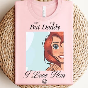 May include: A pale pink t-shirt featuring the text "But Daddy I Love Him" and a cartoon illustration of a character with red hair and blue eyes. The shirt is folded on a woven mat.