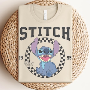 May include: Beige t-shirt featuring a graphic of Stitch from Lilo & Stitch. The design includes the word "STITCH" in dark gray block letters, the year "1995", and a checkered circle around Stitch. The shirt is folded on a woven surface.