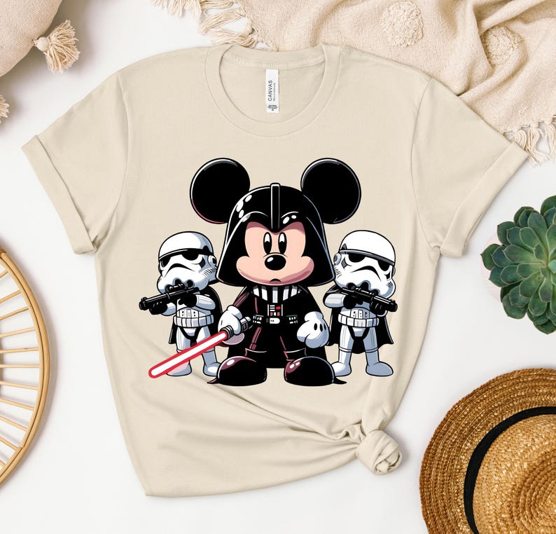 K&ouml;nnte beinhalten: Beiges T-Shirt mit einer Cartoon-Illustration von Mickey Mouse als Darth Vader, der ein rotes Lichtschwert h&auml;lt, flankiert von zwei Sturmtruppen. Das Design ist eine spielerische Mischung aus Disney- und Star-Wars-Themen.