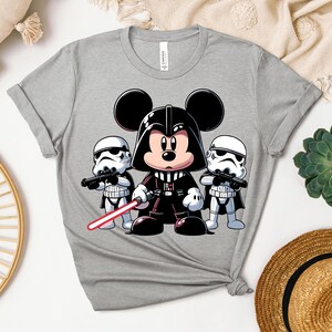 K&ouml;nnte beinhalten: Graues T-Shirt mit einer Cartoon-Illustration von Mickey Mouse als Darth Vader, der ein rotes Lichtschwert h&auml;lt, flankiert von zwei Sturmtruppen. Das Design ist im Cartoon-Stil gehalten, mit kr&auml;ftigen Linien und leuchtenden Farben. Das T-Shirt hat einen Rundhalsausschnitt und kurze &Auml;rmel.