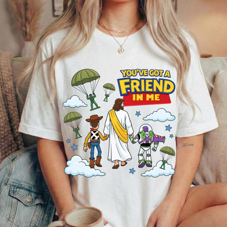Peut inclure: Un t-shirt blanc avec une impression graphique repr&eacute;sentant une figure centrale en robe blanche tenant la main d'une poup&eacute;e cow-boy et d'une poup&eacute;e ranger de l'espace. Le motif inclut des soldats verts avec des parachutes, des &eacute;toiles bleues, des nuages blancs et le texte YOU'VE GOT A FRIEND IN ME.