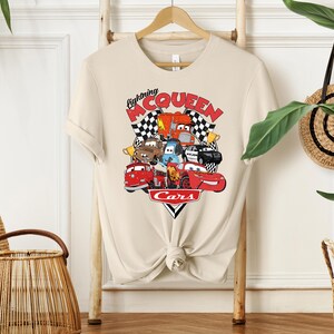 May include: Beige t-shirt featuring a colorful graphic of characters from the movie "Cars." The design includes the text "Lightning McQueen" and "Cars" with a checkered flag background. The shirt is hanging on a wooden hanger.