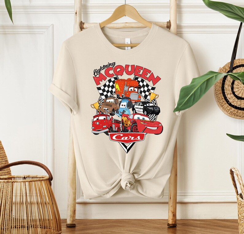 May include: Beige t-shirt featuring a colorful graphic of characters from the movie "Cars." The design includes the text "Lightning McQueen" and "Cars" with a checkered flag background. The shirt is hanging on a wooden hanger.