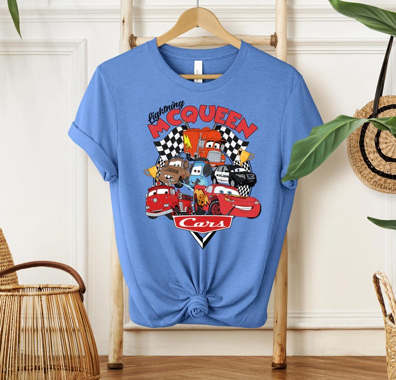 May include: A light blue t-shirt featuring a graphic of characters from the animated movie "Cars." The design includes the text "Lightning McQueen" and "Cars" with images of various cars, checkered flags, and a red race car.