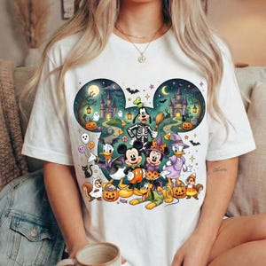 K&ouml;nnte beinhalten: Ein wei&szlig;es T-Shirt mit einer gro&szlig;en Grafik in Form von Mickey-Mouse-Ohren, die eine Halloween-Szene zeigt. Das Design umfasst Zeichentrickfiguren in Kost&uuml;men, ein gruseliges Schloss, K&uuml;rbisse, Flederm&auml;use, Geister und Sterne am Nachthimmel.