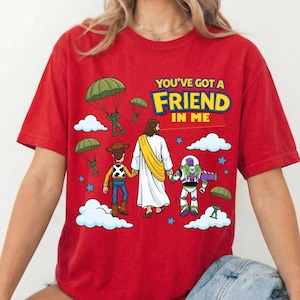 Peut inclure: Un t-shirt rouge &agrave; manches courtes avec un motif repr&eacute;sentant une figure centrale en robe blanche tenant la main d'une poup&eacute;e cow-boy et d'une poup&eacute;e ranger de l'espace. Le design inclut des soldats verts en parachute, des nuages blancs, des &eacute;toiles bleues et le texte "YOU'VE GOT A FRIEND IN ME" en lettres jaunes et bleues.