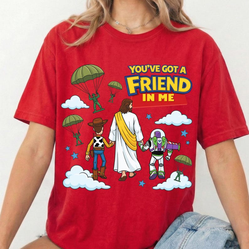 Peut inclure: Un t-shirt rouge &agrave; manches courtes avec un motif repr&eacute;sentant une figure centrale en robe blanche tenant la main d'une poup&eacute;e cow-boy et d'une poup&eacute;e ranger de l'espace. Le design inclut des soldats verts en parachute, des nuages blancs, des &eacute;toiles bleues et le texte "YOU'VE GOT A FRIEND IN ME" en lettres jaunes et bleues.
