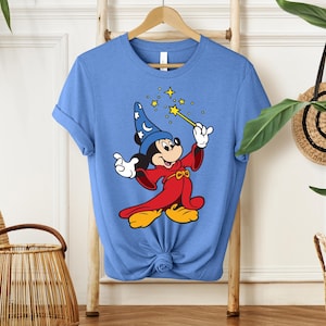 May include: A light blue t-shirt featuring a cartoon Mickey Mouse dressed as a wizard. Mickey is wearing a blue wizard hat with white stars, a red robe, and yellow shoes. He is holding a gold wand with stars.