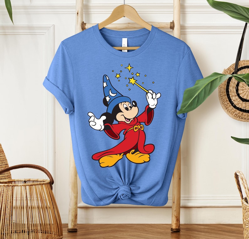 May include: A light blue t-shirt featuring a cartoon Mickey Mouse dressed as a wizard. Mickey is wearing a blue wizard hat with white stars, a red robe, and yellow shoes. He is holding a gold wand with stars.