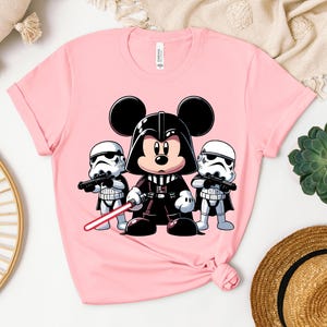 K&ouml;nnte beinhalten: Hellrosa T-Shirt mit einer Cartoon-Illustration von Mickey Mouse als Darth Vader, der ein rotes Lichtschwert h&auml;lt, flankiert von zwei Sturmtruppen. Ein verspieltes Mashup von Disney und Star Wars.