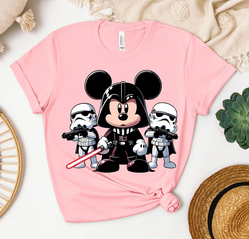 K&ouml;nnte beinhalten: Hellrosa T-Shirt mit einer Cartoon-Illustration von Mickey Mouse als Darth Vader, der ein rotes Lichtschwert h&auml;lt, flankiert von zwei Sturmtruppen. Ein verspieltes Mashup von Disney und Star Wars.