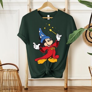 May include: Dark green t-shirt featuring a cartoon character in a red robe and blue wizard hat, holding a yellow wand with stars. The character has yellow shoes and white gloves. The shirt is hanging on a wooden hanger.