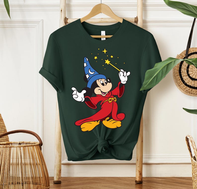May include: Dark green t-shirt featuring a cartoon character in a red robe and blue wizard hat, holding a yellow wand with stars. The character has yellow shoes and white gloves. The shirt is hanging on a wooden hanger.