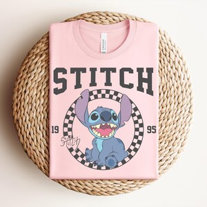 May include: A folded pink t-shirt featuring a graphic of the character Stitch from the movie Lilo & Stitch. The design includes the word "STITCH" in large, dark gray letters, the year "1995", and a checkered circle around Stitch.