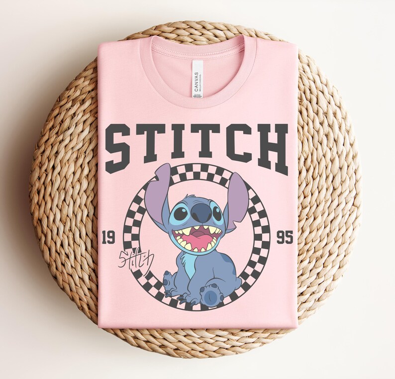 May include: A folded pink t-shirt featuring a graphic of the character Stitch from the movie Lilo & Stitch. The design includes the word "STITCH" in large, dark gray letters, the year "1995", and a checkered circle around Stitch.