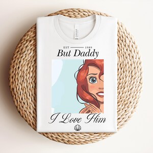 May include: White t-shirt featuring the text "But Daddy I Love Him" and a cartoon illustration of a character with red hair and blue eyes. The shirt is folded on a woven mat.