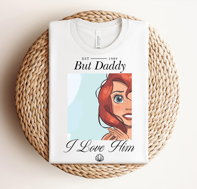 May include: White t-shirt featuring the text "But Daddy I Love Him" and a cartoon illustration of a character with red hair and blue eyes. The shirt is folded on a woven mat.