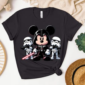 K&ouml;nnte beinhalten: Schwarzes T-Shirt mit einer Cartoon-Illustration von Mickey Mouse als Darth Vader, der ein rotes Lichtschwert h&auml;lt, flankiert von zwei Sturmtruppen. Ein lustiges Design, das Disney- und Star Wars-Themen kombiniert.