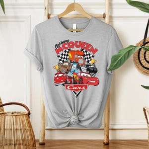 May include: A light gray t-shirt featuring a colorful graphic of characters from the movie Cars. The graphic includes the text "Lightning McQueen" and "Cars" with checkered flags. The shirt is hanging on a wooden rack.