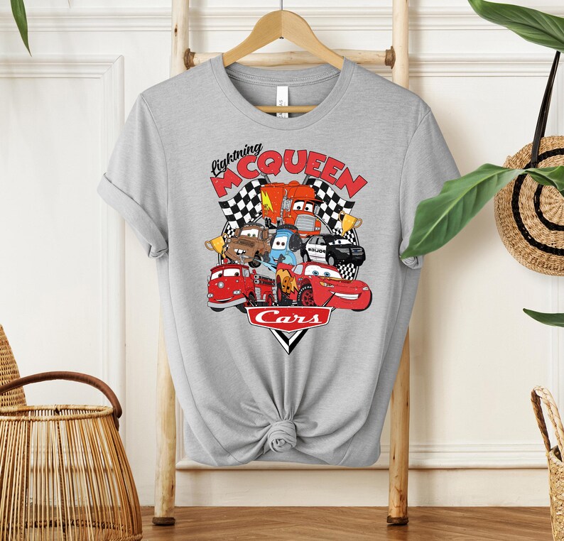 May include: A light gray t-shirt featuring a colorful graphic of characters from the movie Cars. The graphic includes the text "Lightning McQueen" and "Cars" with checkered flags. The shirt is hanging on a wooden rack.