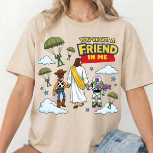 Peut inclure: Un t-shirt beige avec une impression graphique repr&eacute;sentant un cow-boy, un ranger de l'espace et une silhouette en robe blanche avec une &eacute;charpe jaune marchant ensemble. Le texte indique YOU'VE GOT A FRIEND IN ME, avec des soldats verts en parachute.