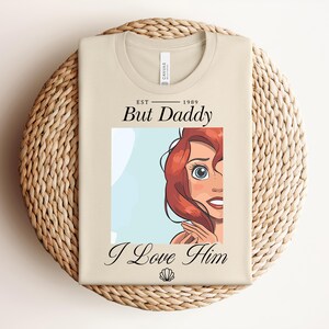 May include: A beige t-shirt featuring the text "EST 1989 But Daddy I Love Him" and a cartoon illustration of a character with red hair and blue eyes. The shirt is folded on a woven mat.