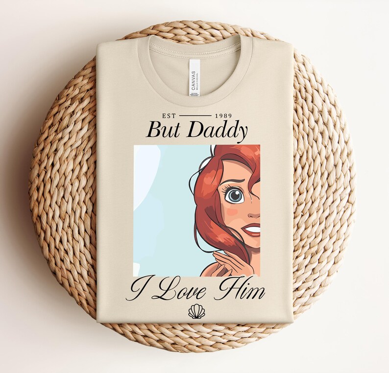 May include: A beige t-shirt featuring the text "EST 1989 But Daddy I Love Him" and a cartoon illustration of a character with red hair and blue eyes. The shirt is folded on a woven mat.